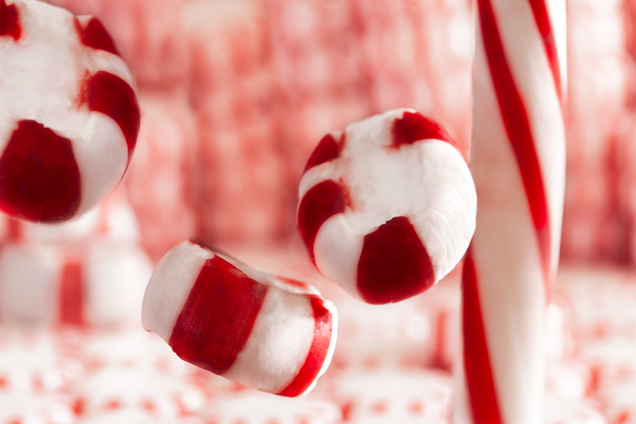 This Brian Charles Steel photograph is composed of a peppermint stick, oval shaped peppermints and circular peppermints. The circular peppermints compose the background in stacks and are out of focus. On the right side there is peppermint stick going straight down from top to bottom. In front of the stick was an oval shaped peppermint floating near the top center. Below the floating mint was another floating oval shaped mint. There was another floating mint in the top left corner.  